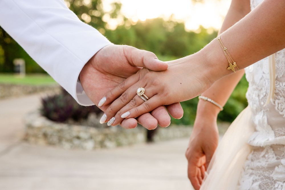 Close-up of hands wearing wedding rings holding each other against a blurred outdoor background.