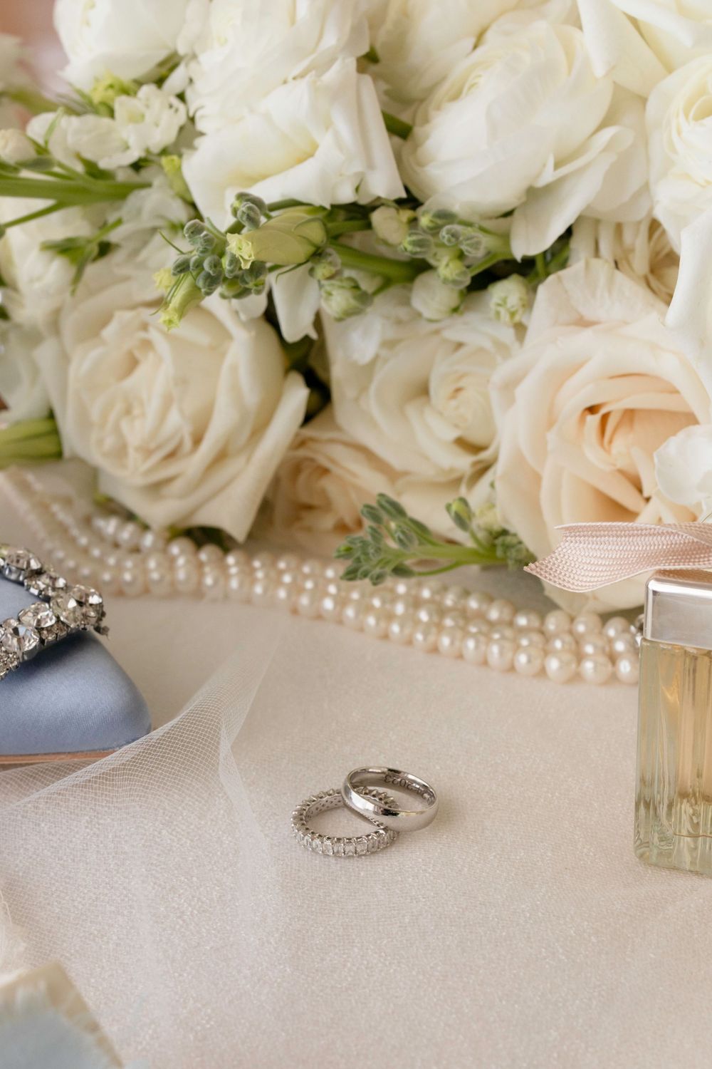 Elegant wedding details featuring white roses, pearl necklaces, and diamond rings on a cream-colored surface.