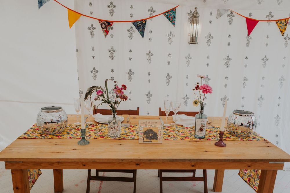 Rustic wooden wedding table decorated with colorful bunting, floral runner, and wildflower centerpieces.