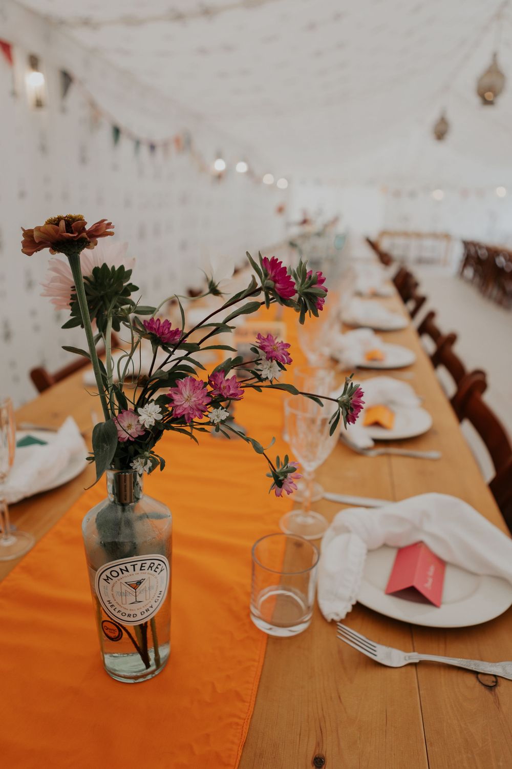 Elegant wedding reception table with orange runner, wildflowers in gin bottle vase, and white place settings.