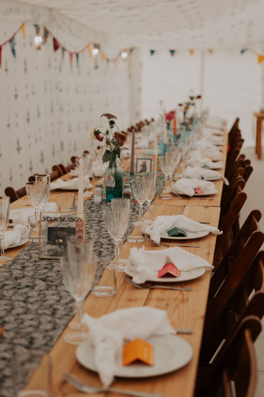 Long wedding banquet table with crystal glasses, floral centerpieces, and colorful bunting in a marquee.