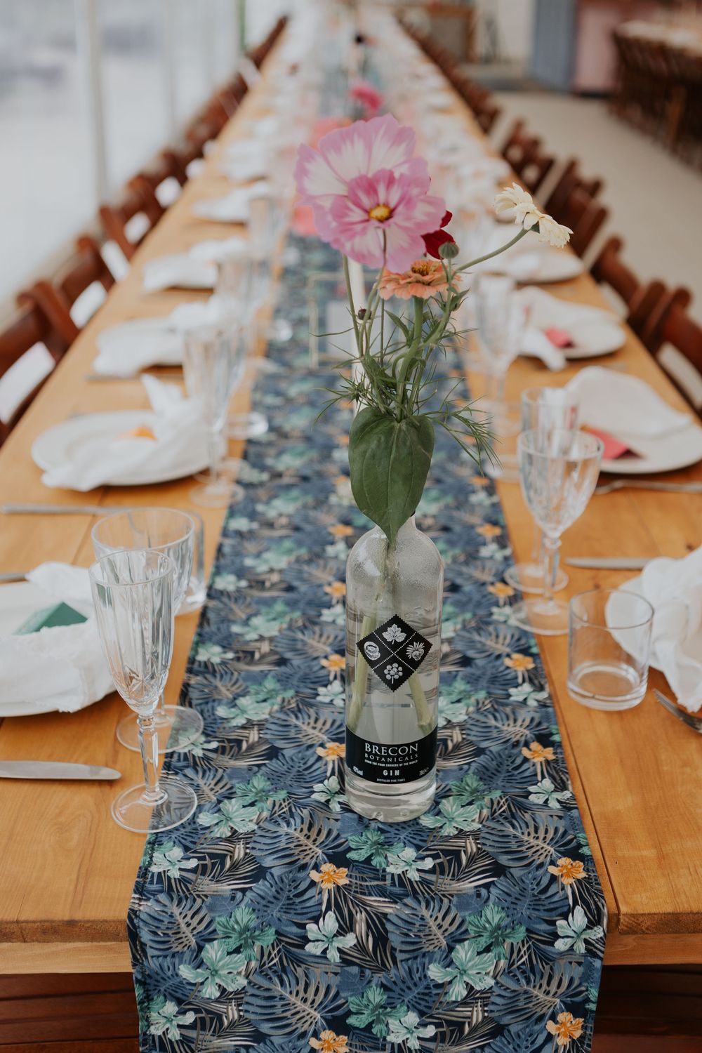 Wedding table with dark floral runner, wildflowers in Brecon gin bottle vase, and crystal glassware.