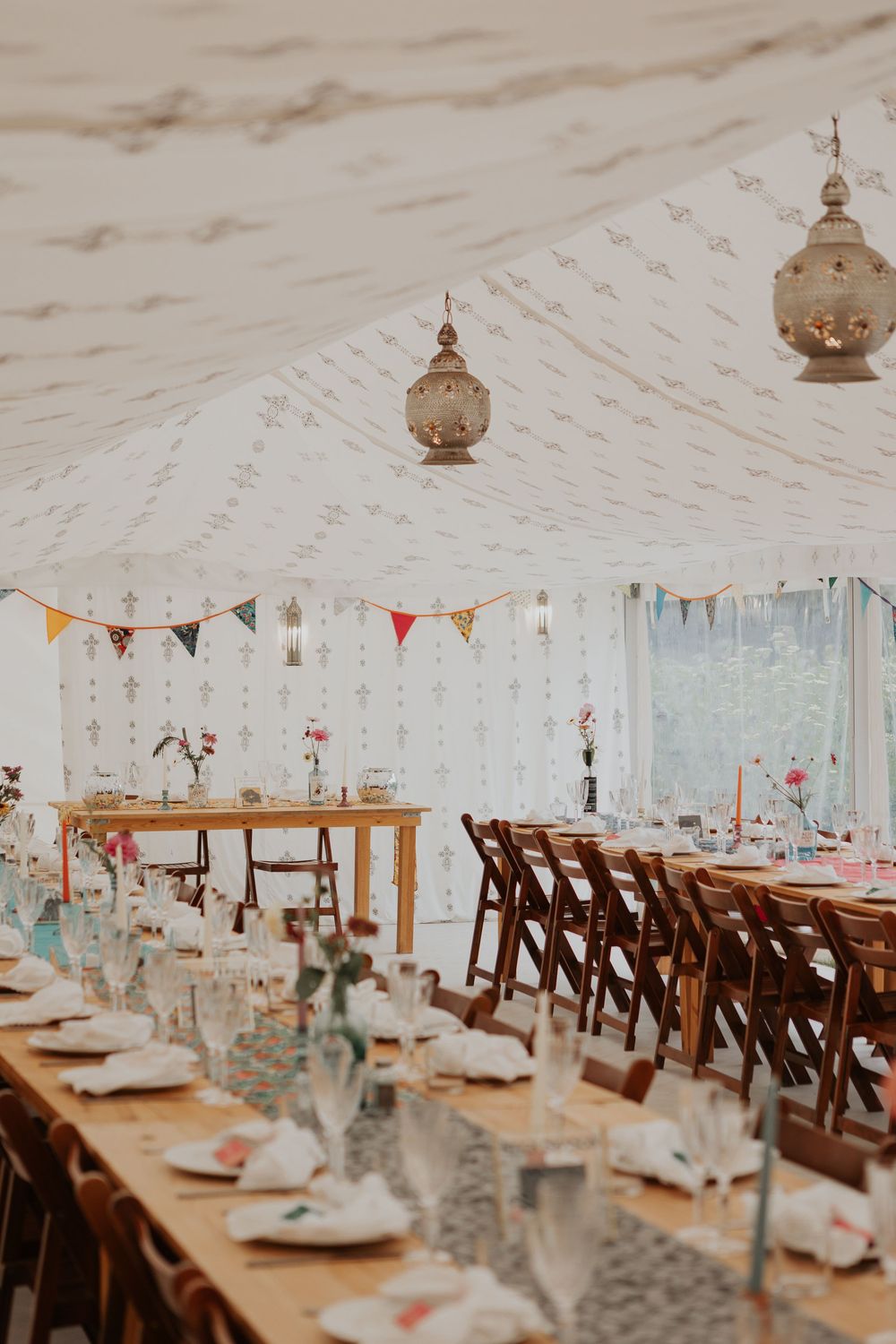 Wedding marquee interior with long dining tables, Moroccan hanging lanterns, colorful bunting, and wooden chairs.