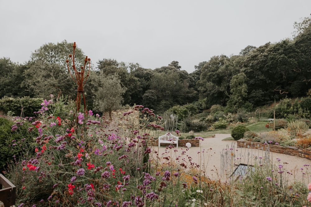 Colorful wildflower garden with pink and purple blooms, lush green trees, and a rustic outdoor wedding venue in the background.