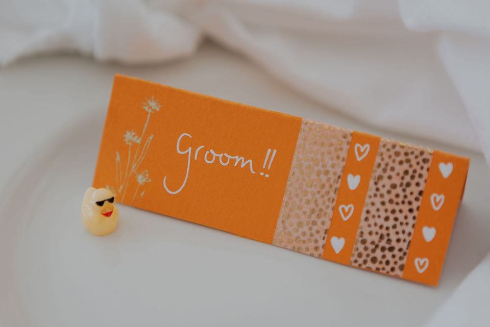Orange handmade groom place card with hearts and floral design beside small duck figurine.