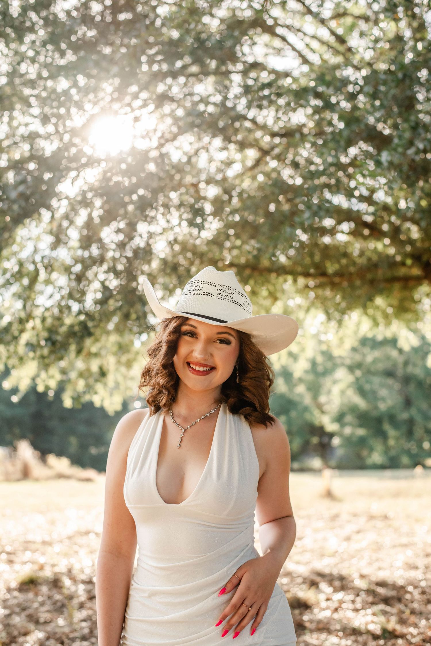 Person in a white sundress and floppy hat sits in a sun-dappled outdoor setting during golden hour.