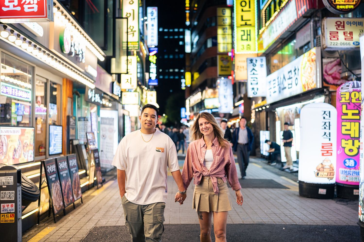 Two people walk together at night through a vibrant street with colorful Korean signs and storefronts in Seoul.