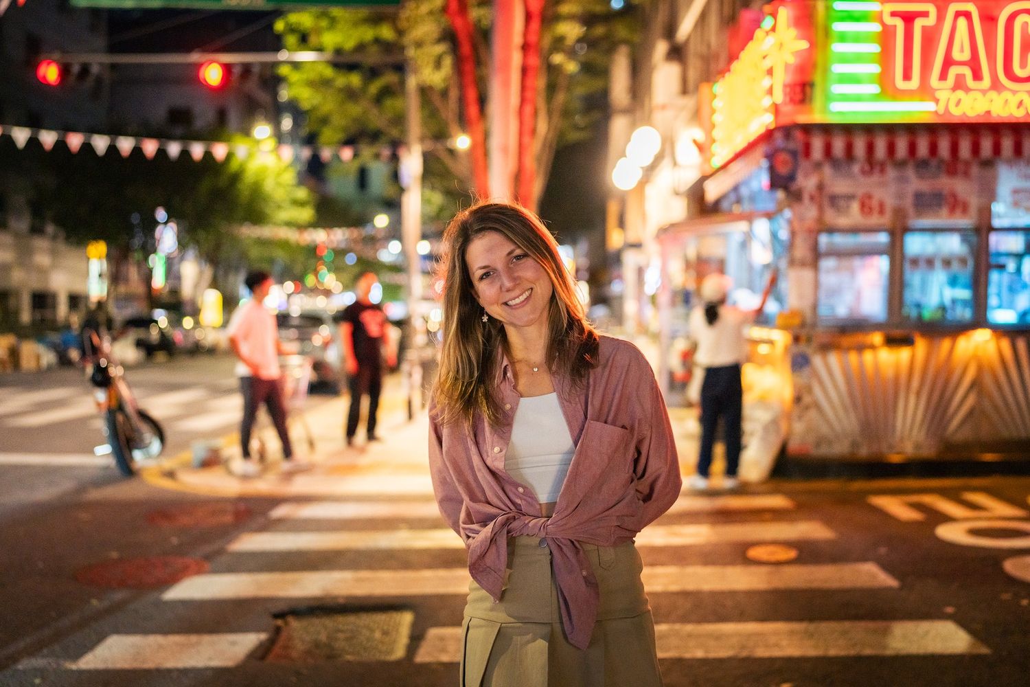 A person stands smiling on a crosswalk at night with colorful neon signs and city lights in the background.
