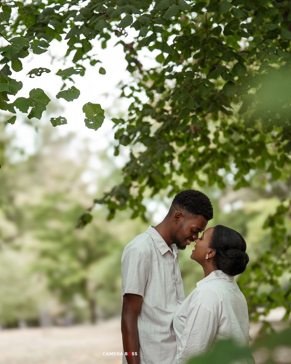 A romantic couple shares an intimate moment under green tree leaves in matching white outfits.