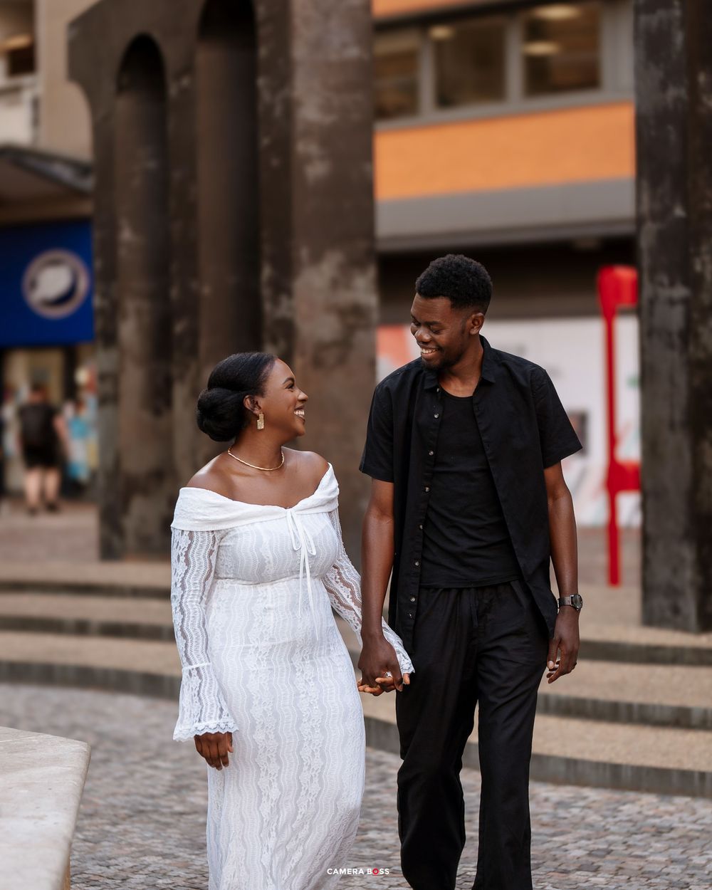 A couple in formal attire walks together on a cobblestone street during an engagement photoshoot.
