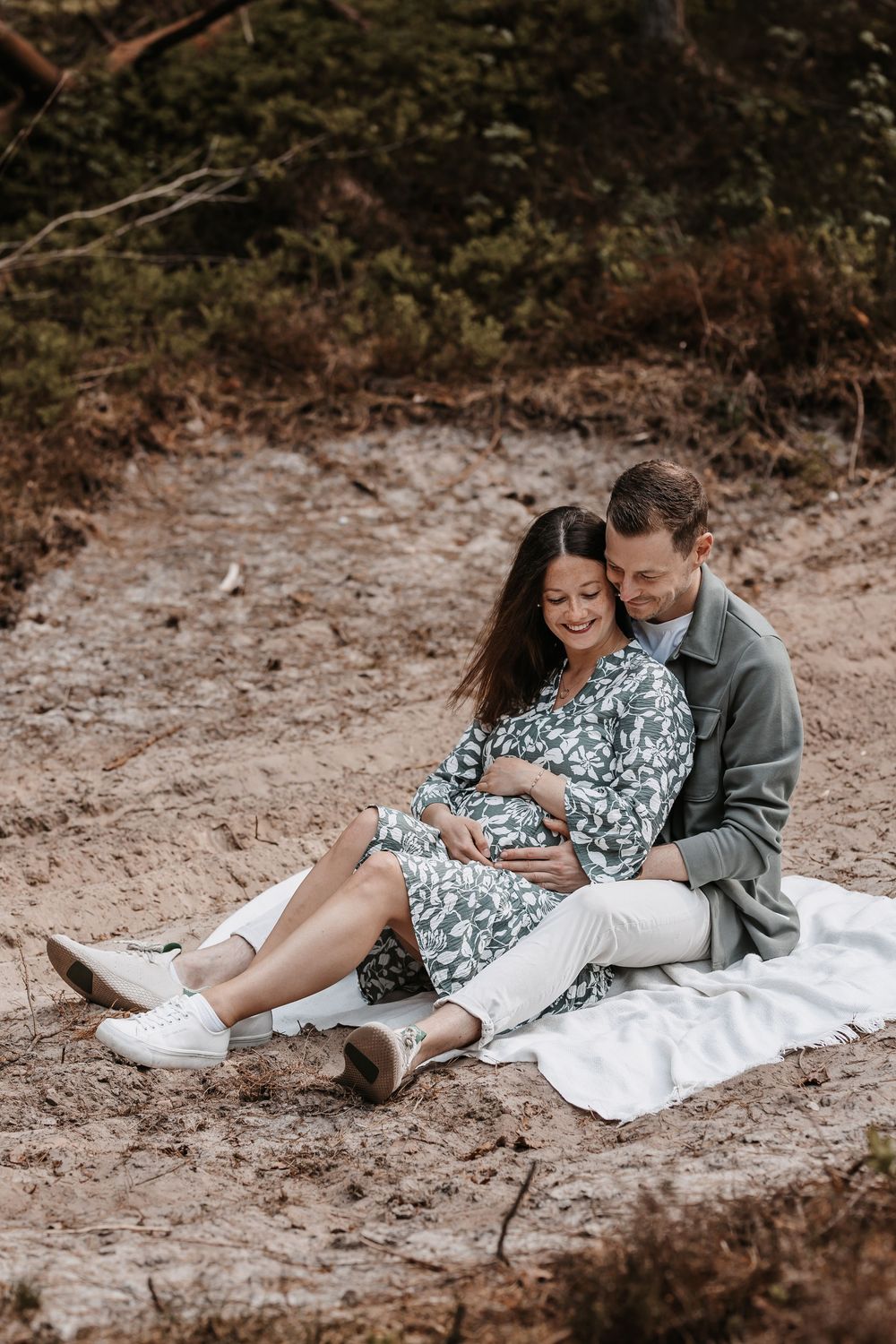 Couple lounges on white blanket during forest photoshoot wearing casual attire.