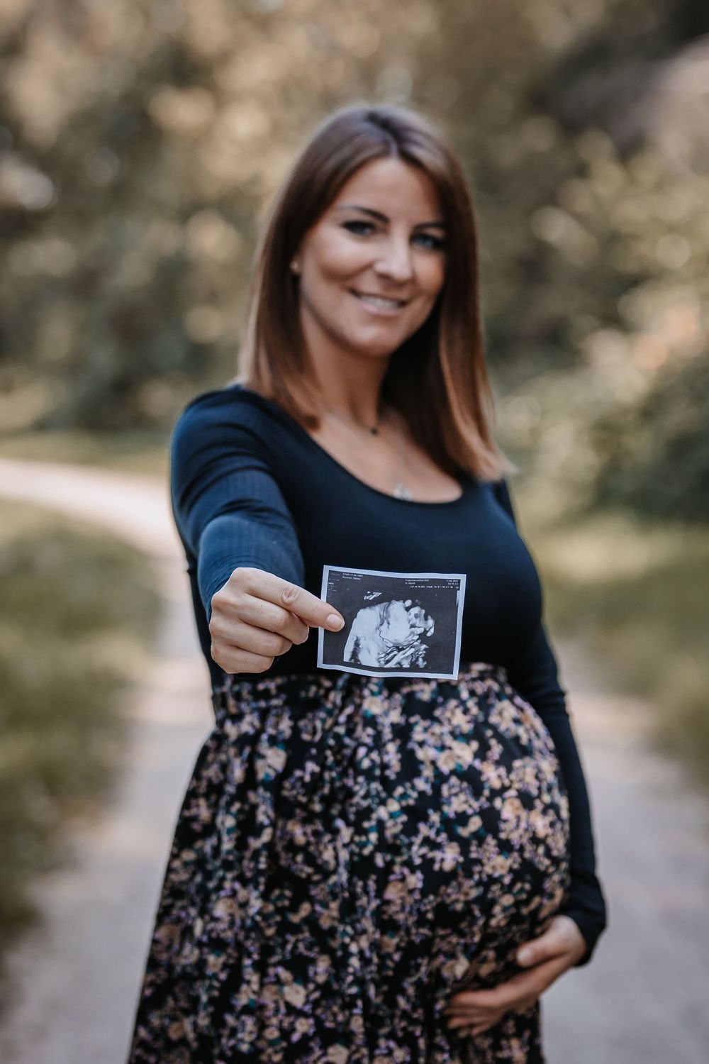 Expectant mother in floral dress holds ultrasound photo while standing on outdoor path.