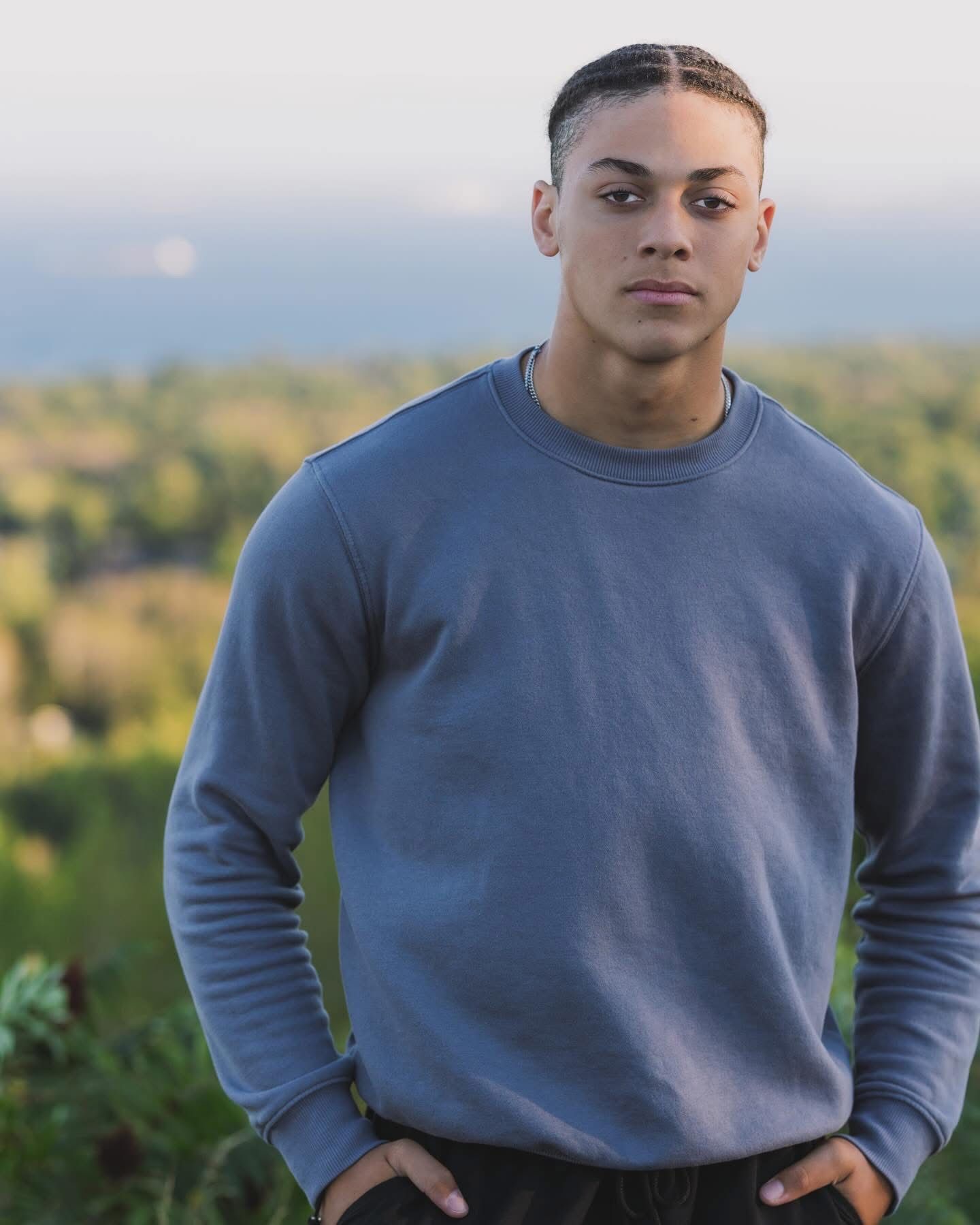 A person wearing a blue-gray sweatshirt poses outdoors against a scenic natural landscape during golden hour.