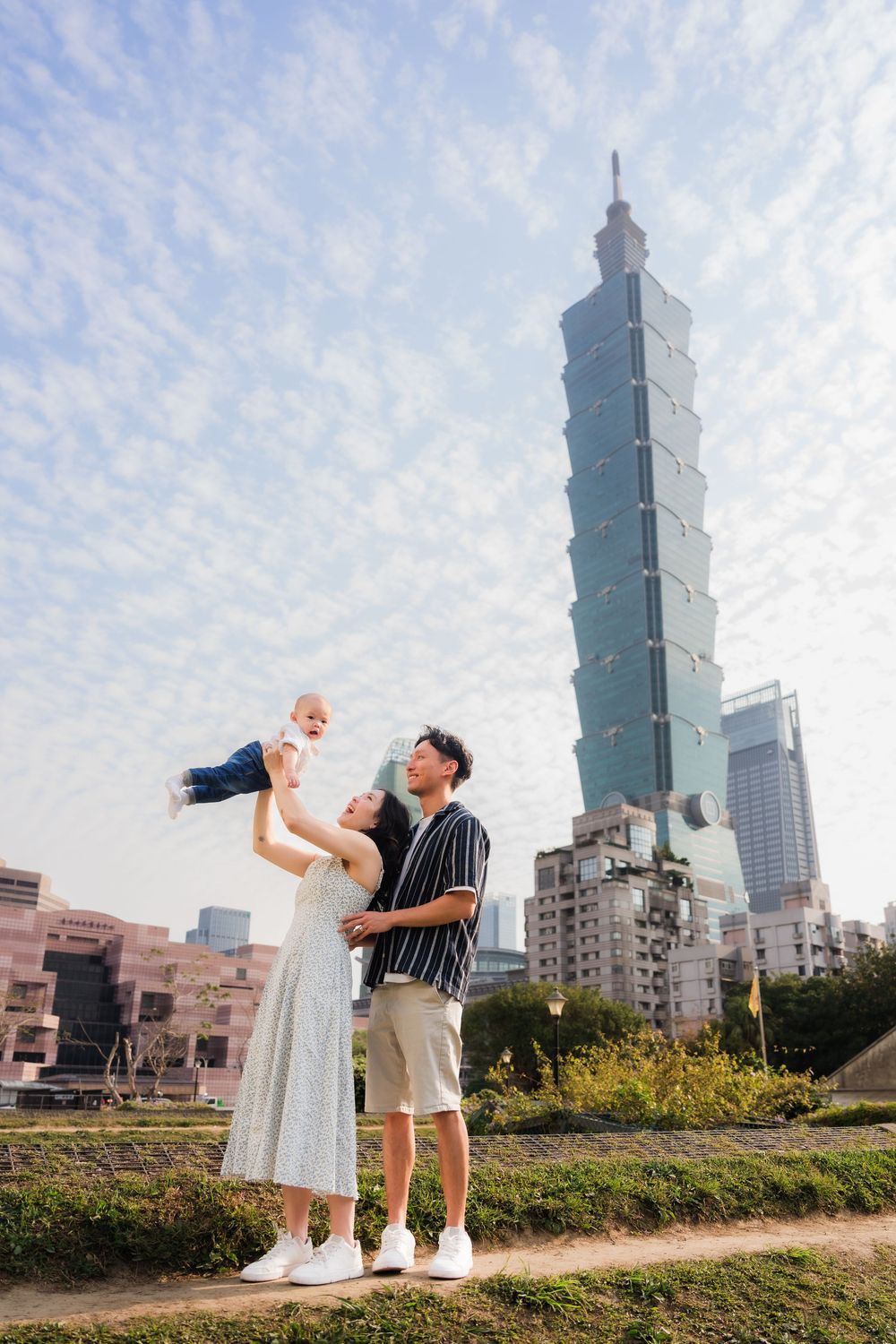 A couple poses playfully against the backdrop of Taipei 101 tower on a sunny day with wispy clouds.