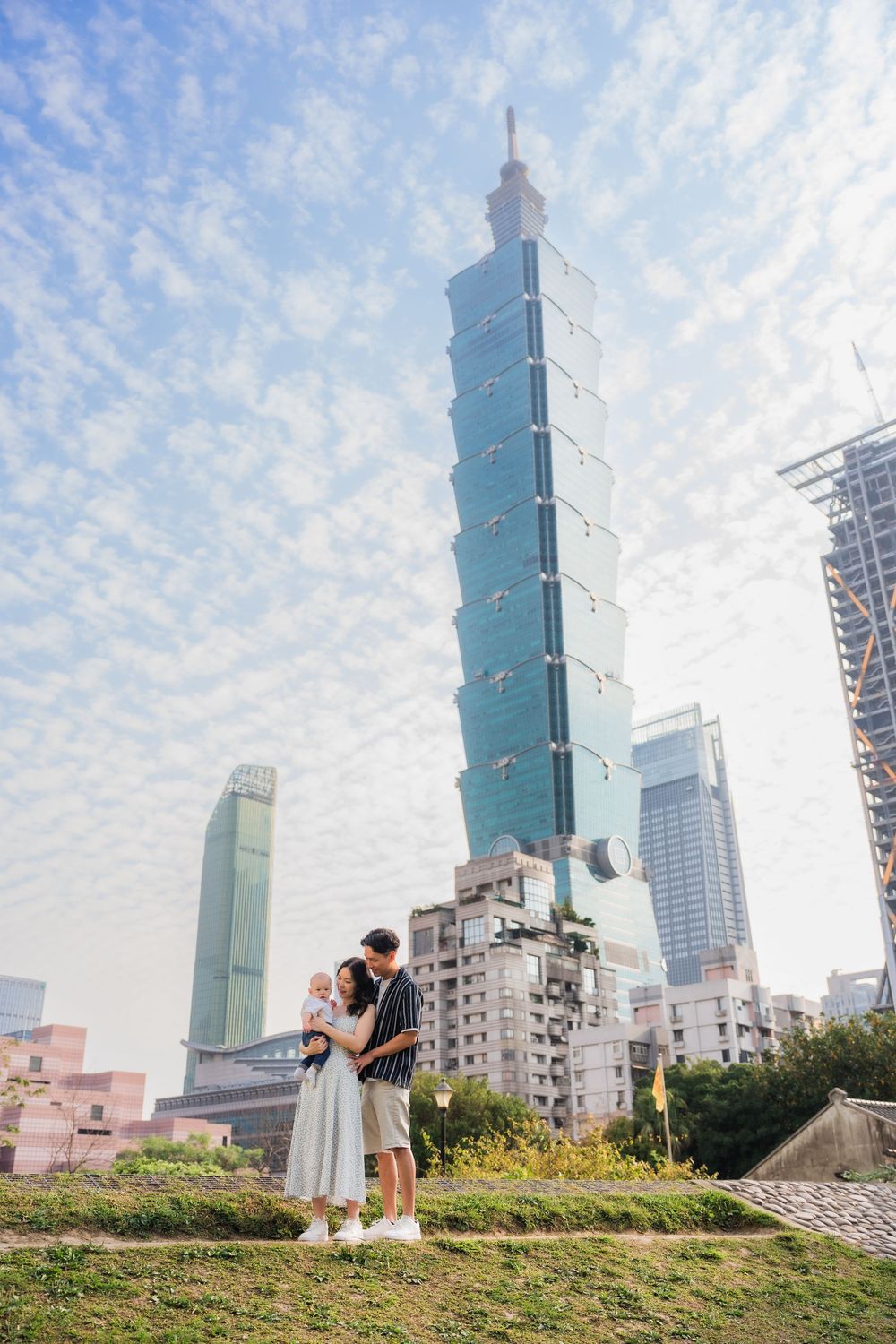 A couple embraces on a grassy hill overlooking Taipei 101 and the city skyline on a beautiful cloudy day.