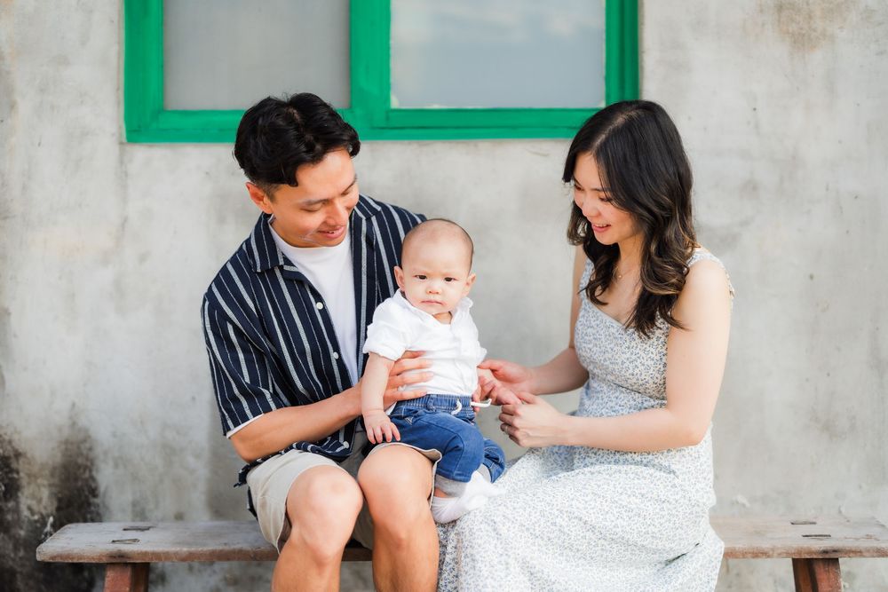 A couple sits on a concrete bench while holding a baby in a white dress.