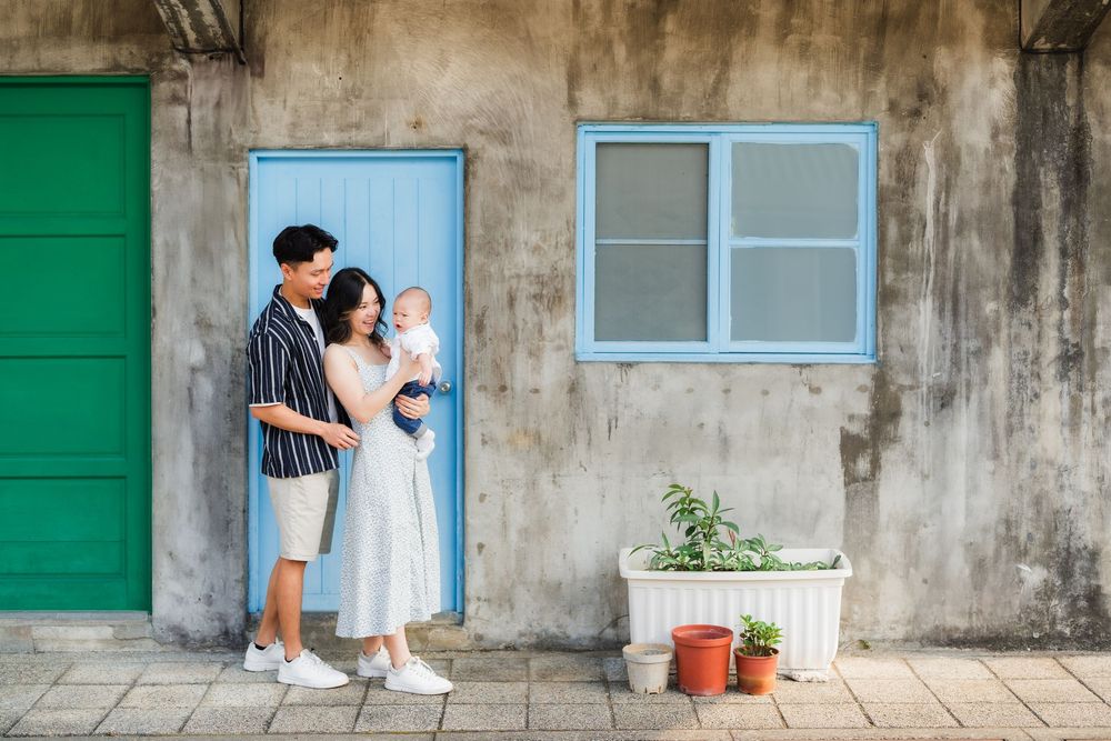A romantic couple in white outfits poses together against a weathered concrete wall with green shutters.