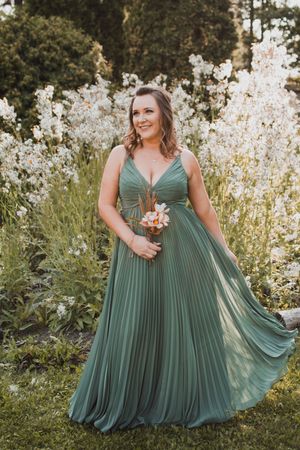 A bridesmaid in a flowing sage green pleated gown poses in front of blooming white flowers at sunset.