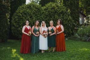 Wedding party in burgundy and green dresses poses together on a grassy lawn with trees in the background.
