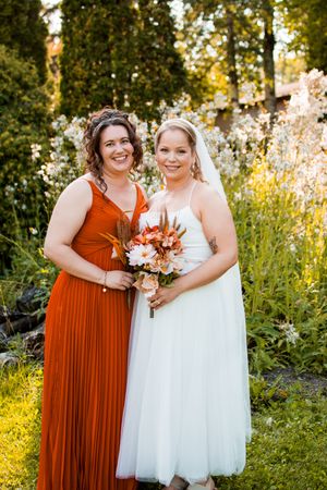 Wedding party members pose in vibrant orange dresses against a backdrop of white flowering plants.