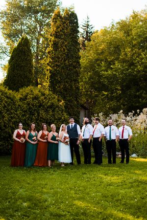Large wedding party in formal attire lines up for group photos on a grassy lawn with trees in background.