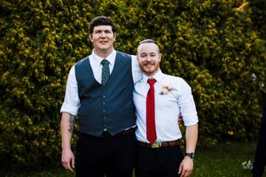 Wedding party members in formal attire posing outdoors with green hedges in the background.