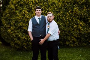 Two people in formal attire pose together in front of lush green hedges at an outdoor event.