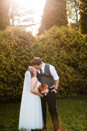 A romantic wedding couple embraces in a garden at sunset with golden light filtering through trees.