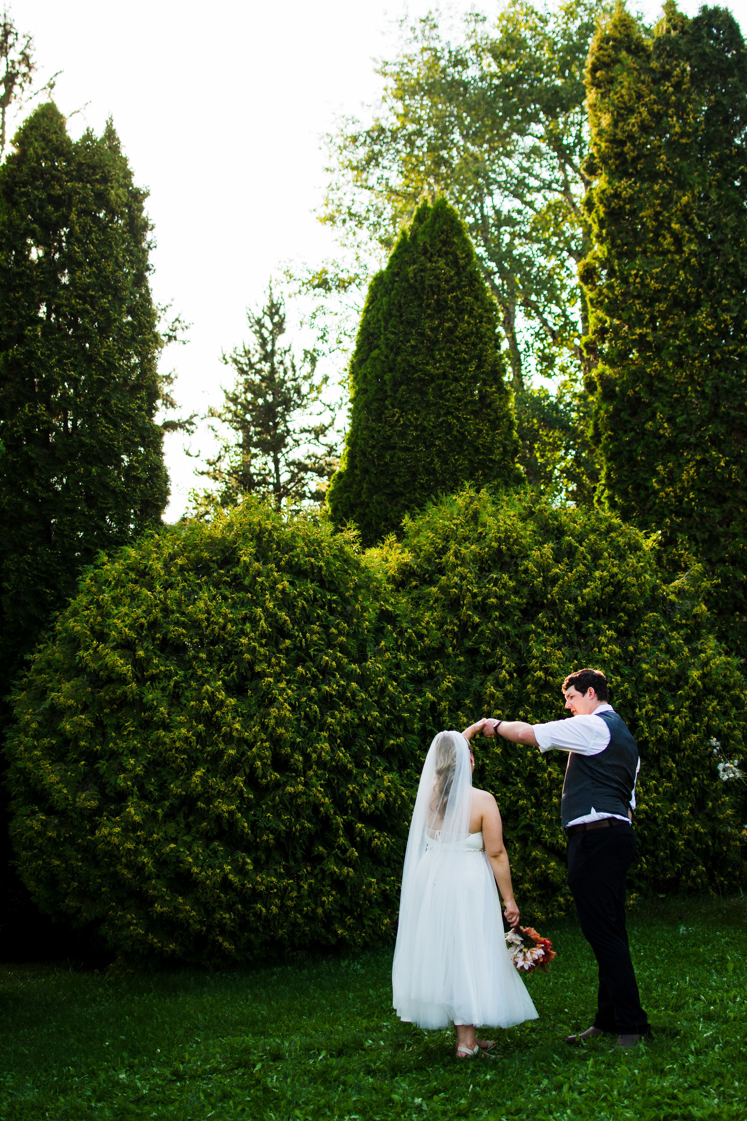 Newlyweds dance together on a grassy lawn surrounded by tall evergreen trees.