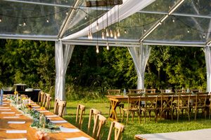 Outdoor wedding reception setup with clear tent, wooden chairs, and elegant table settings.
