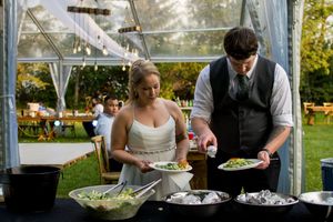 A couple serves food from a buffet line under a clear tent at an outdoor wedding reception.