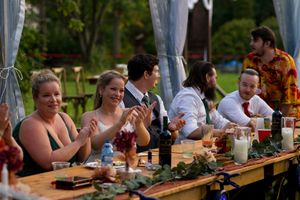 Guests enjoy a casual outdoor dinner party at a long wooden table decorated with greenery under draped fabric.