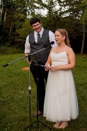 Couple in wedding attire shares a lighthearted moment at a microphone stand outdoors.
