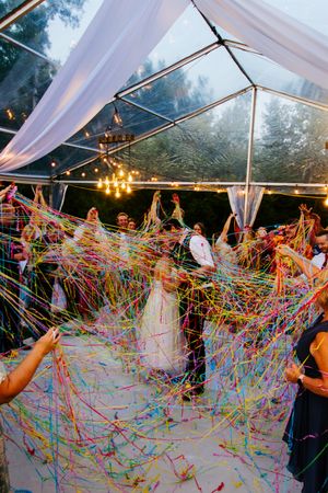 Colorful streamers create a vibrant canopy under a clear wedding tent during a celebration.