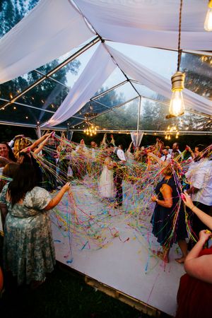 Wedding guests throw colorful streamers under a white tent with hanging Edison bulbs.