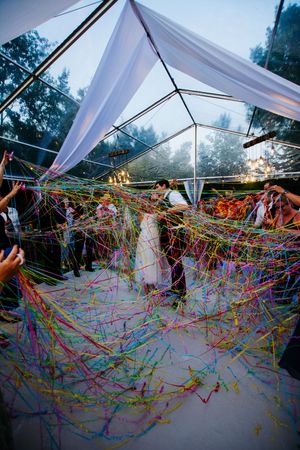 Colorful streamers create a vibrant canopy under a clear tent at a wedding reception.