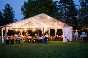 An illuminated white wedding tent glows at dusk with guests dining and celebrating inside.