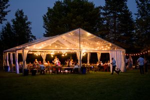Large white wedding tent illuminated at dusk with string lights and guests celebrating inside.