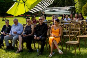 Wedding guests sit on wooden benches under bright yellow umbrellas during an outdoor ceremony.
