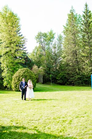 Wedding ceremony taking place on a sunlit lawn surrounded by tall evergreen trees.
