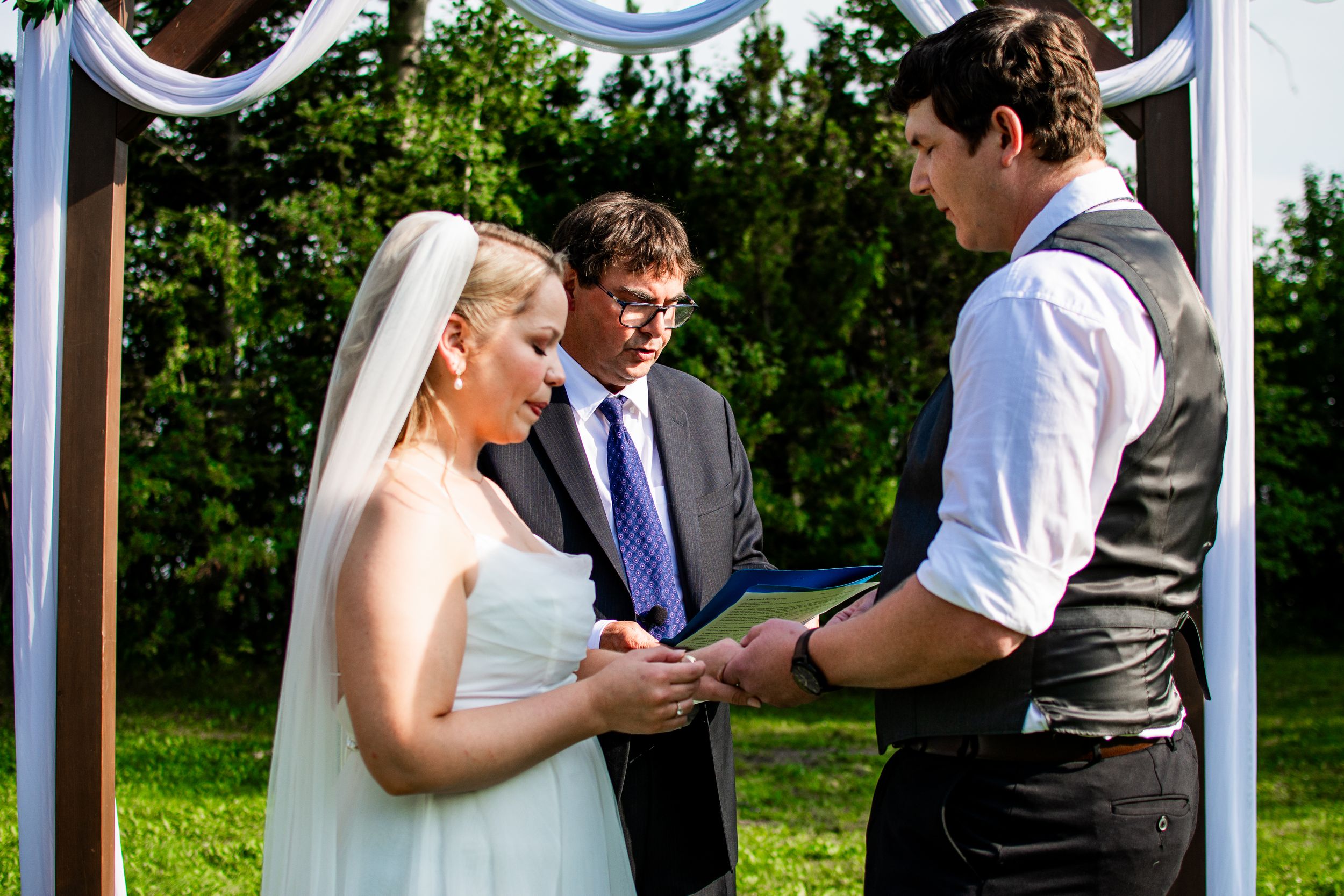 Wedding ceremony takes place under a white arch decorated with flowers in a garden setting.