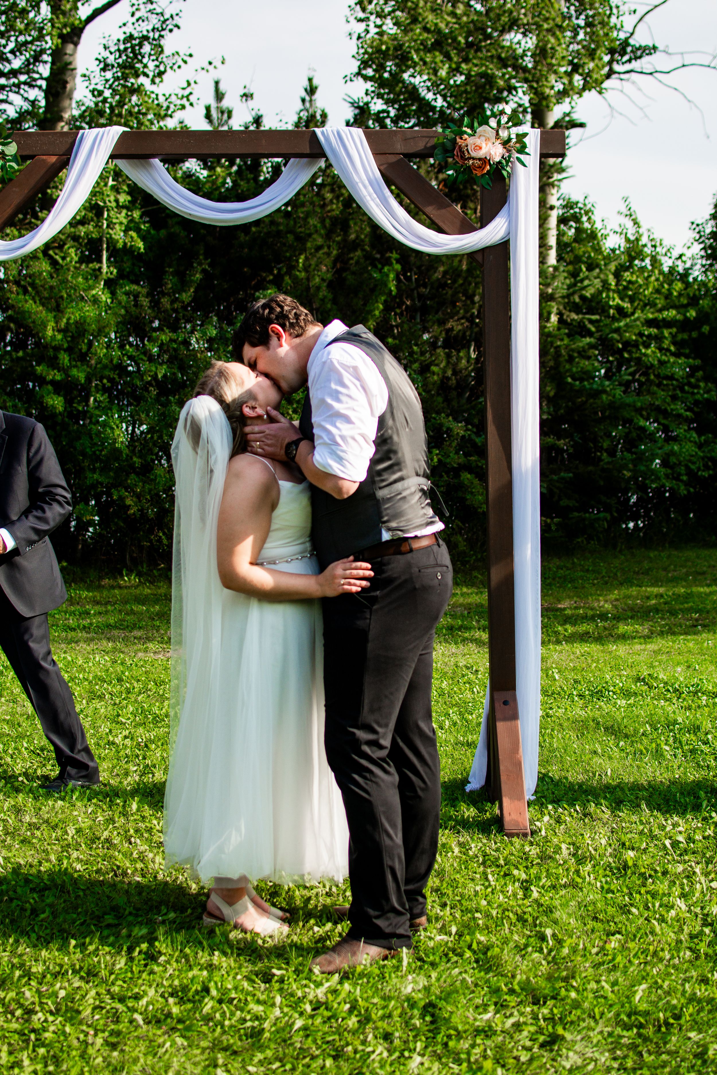 Couple shares their first kiss under a white wedding arch during outdoor ceremony.