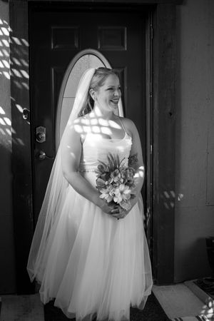 A bride in a white tulle gown and veil stands in dappled sunlight by a dark doorway.
