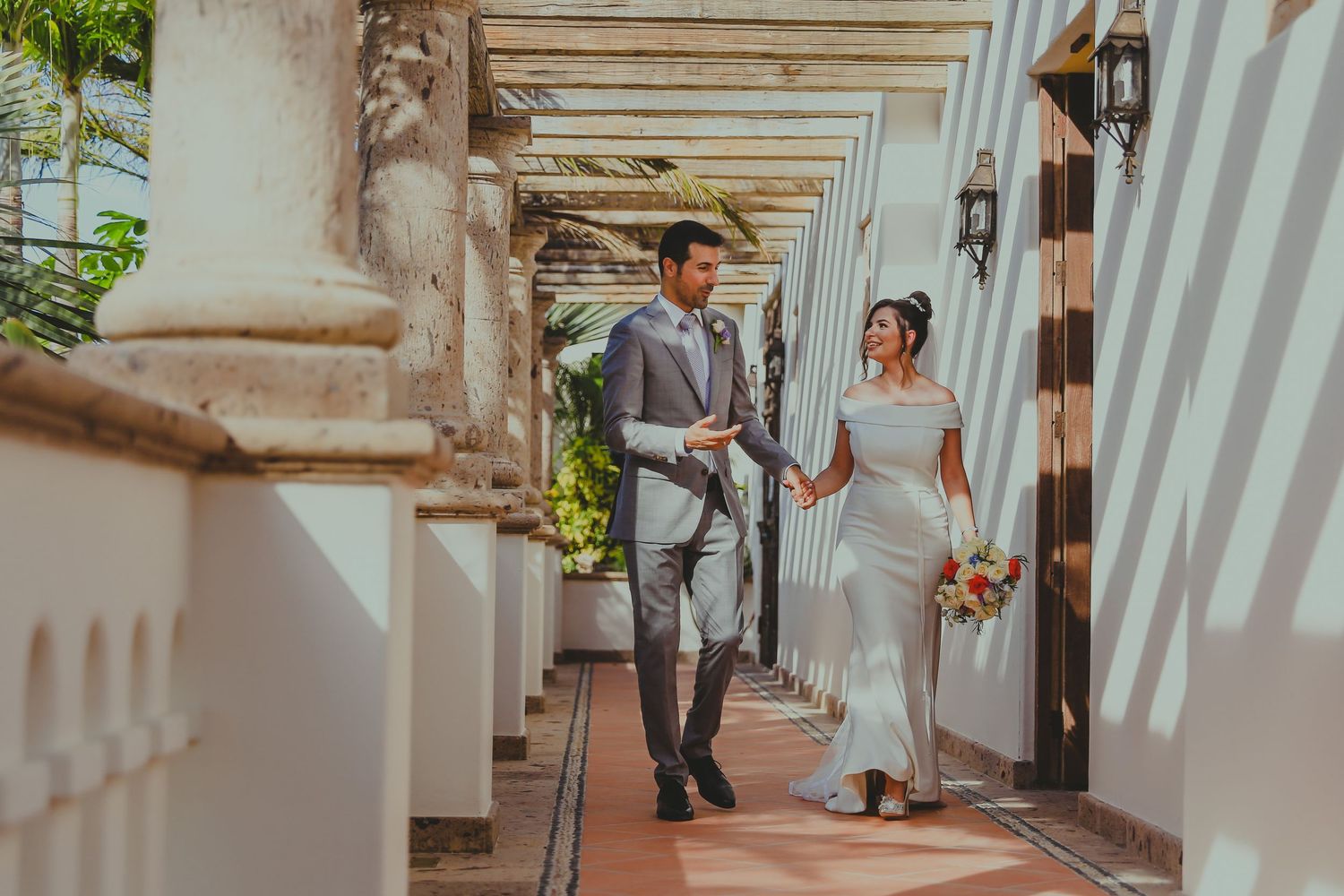 A couple in wedding attire walks through a covered outdoor colonnade with white pillars and sunlit archways.Pedregal Los Cabos Wedding Photo