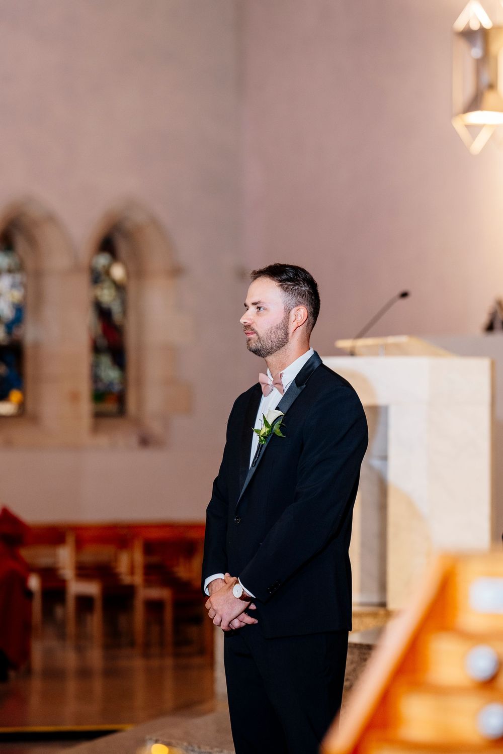 A groom stands solemnly in a black suit at the altar of a church with stained glass windows visible in the background.
