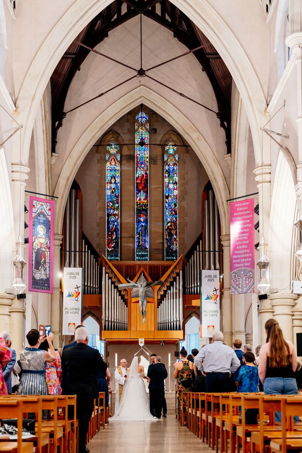 Church interior with stained glass windows, wooden pews, and organ pipes during a wedding ceremony.