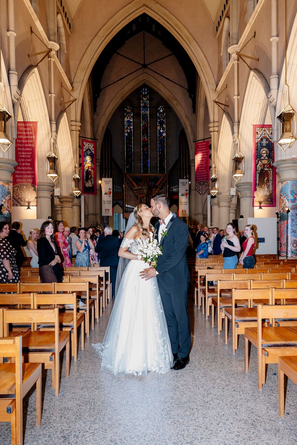 A panoramic view of a church interior with wooden pews and couples in wedding attire spread across the aisle.