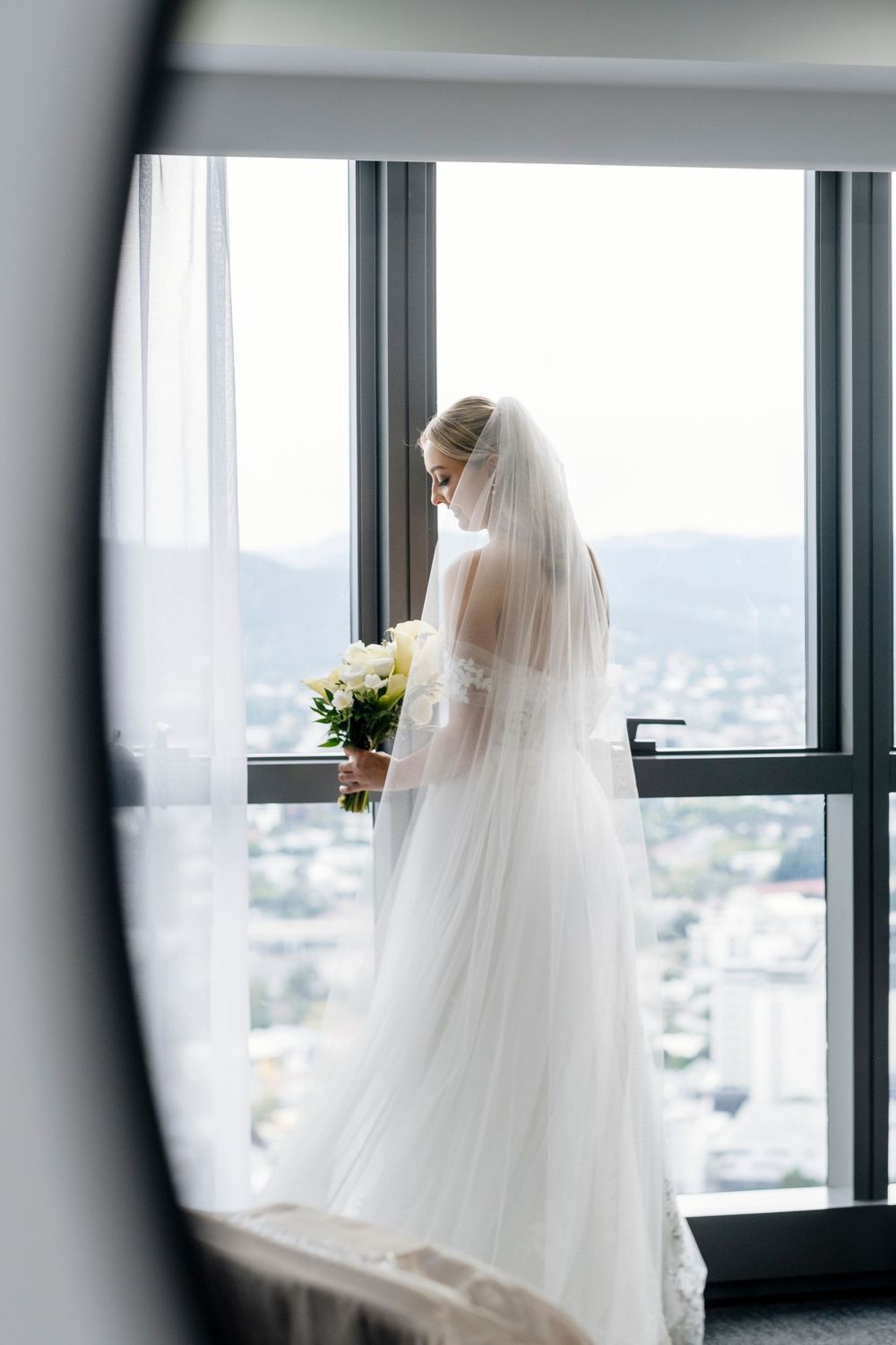 Ethereal silhouette of a bride in a flowing veil stands by large windows overlooking a cityscape.