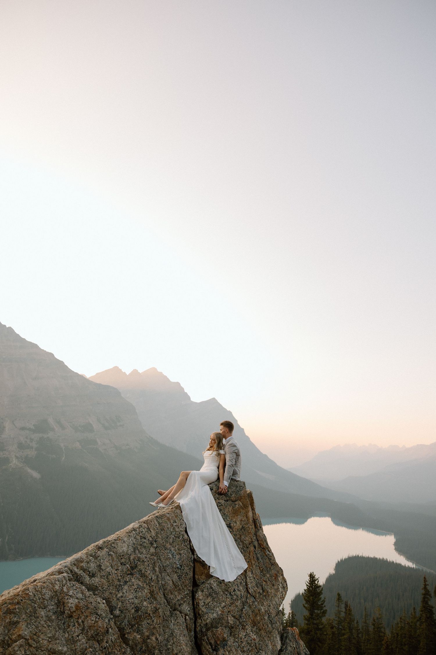 A couple embraces on a mountain peak during sunset with dramatic mountain ranges stretching into the distance.