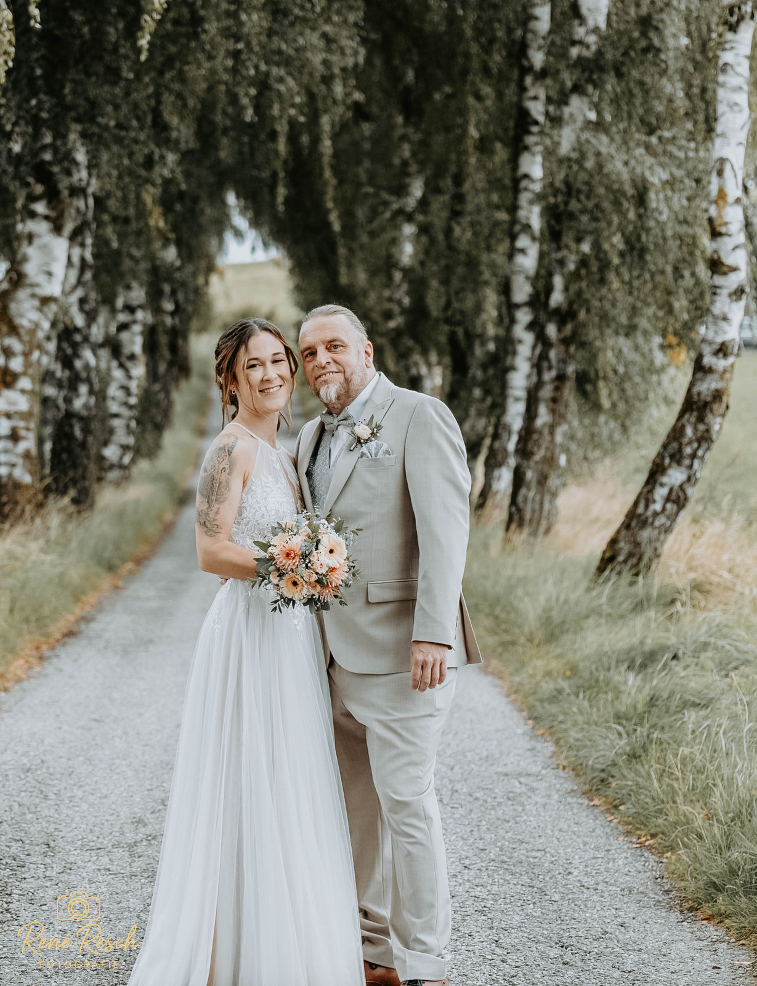 Newlyweds in light gray suit and lace dress pose on tree-lined path.